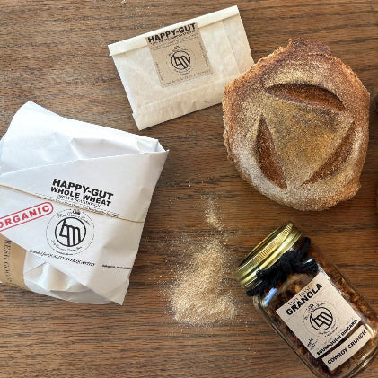 Two packages of 'Happy Gut' on a wooden surface with a loaf of bread and a jar of granola.